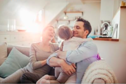 A happy family moment on a couch, with a father hugging his child while the mother smiles. Sunlight fills the cozy room.